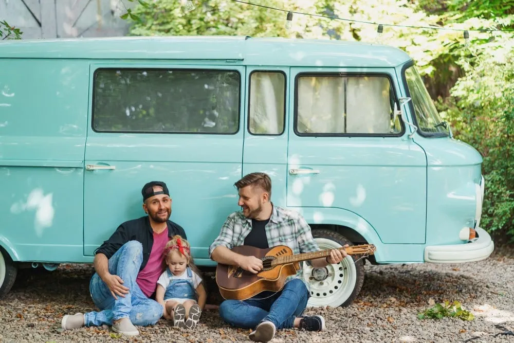 Become a gay parent same-sex couple with child relaxing together outdoors near a van